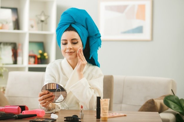 pleased-young-girl-holding-looking-mirror-applying-tone-up-cream-with-sponge-sitting-table-with-makeup-tools-living-room